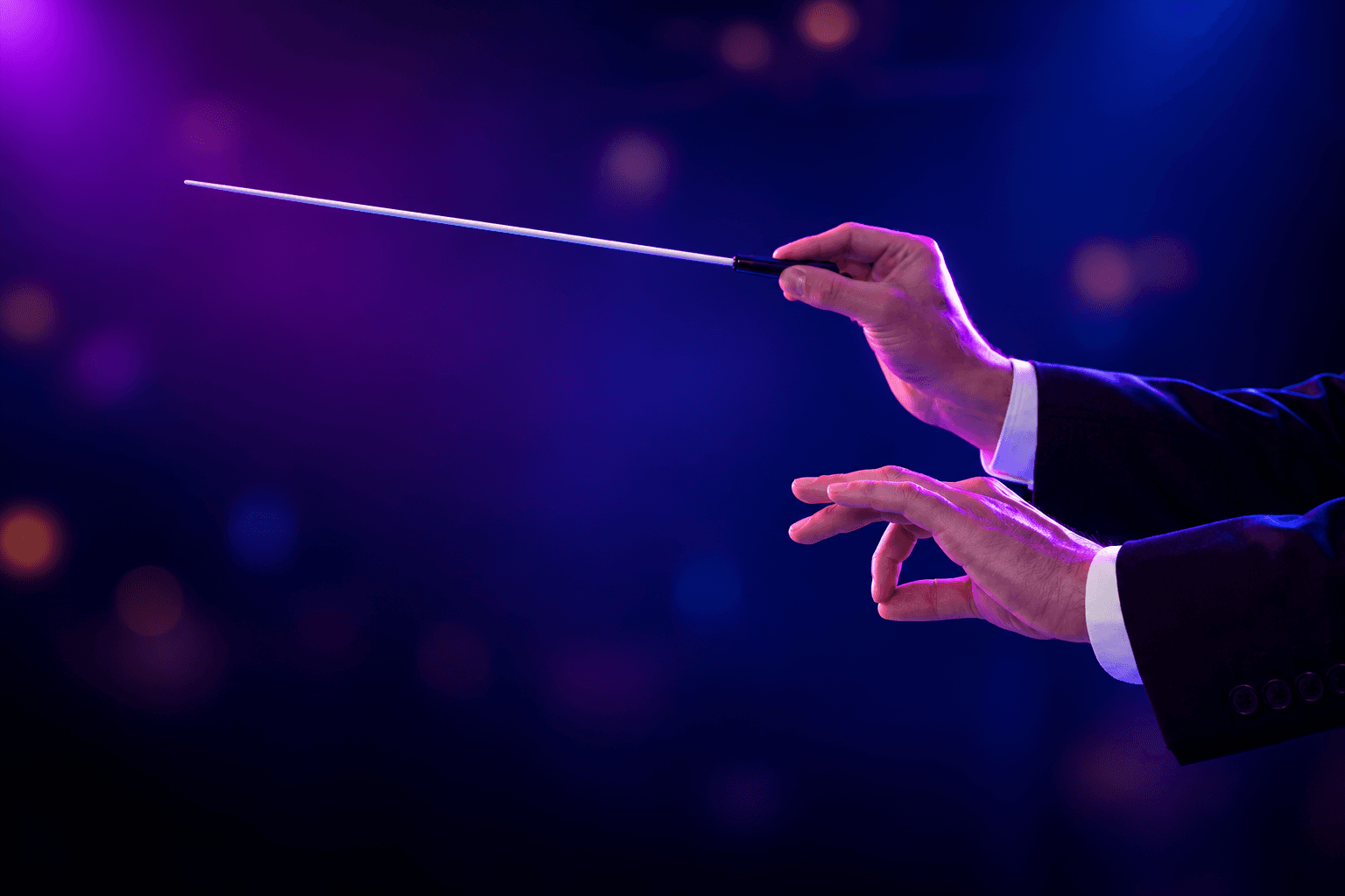 A conductor's hands raised with a baton under purple and blue stage light — the gesture that has to reach every singer in an online choir rehearsal